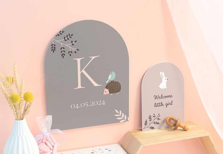 Two personalised arched signs with woodland designs. One grey with a hedgehog, initial, and date; one pink with a rabbit and "Welcome little girl".