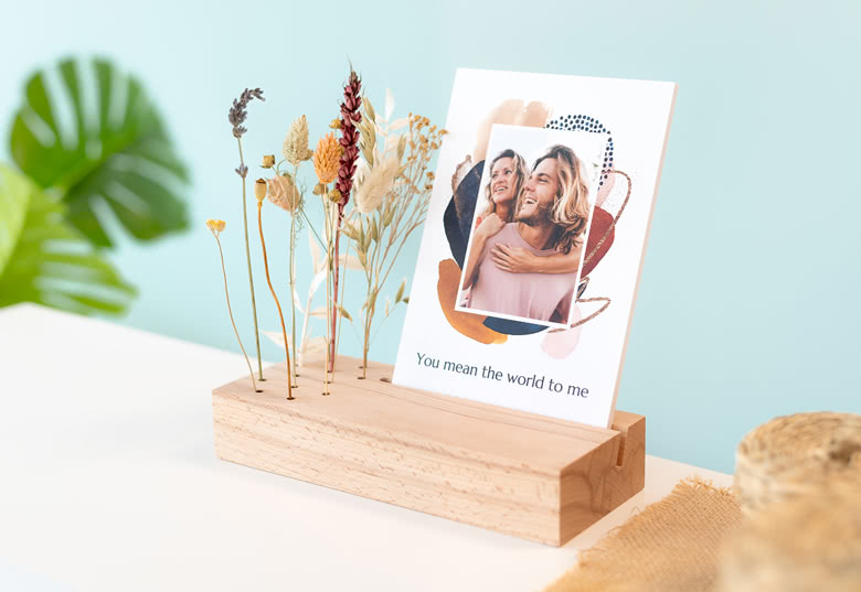 Custom photo refill card with a colourful abstract design, displayed in a wooden stand with dried flowers. Text reads: "You mean the world to me."
